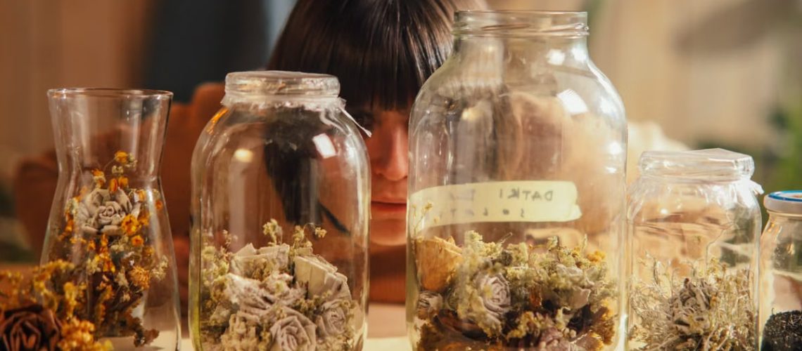 A woman thoughtfully looks at dried flowers arranged in multiple glass containers on a table.
